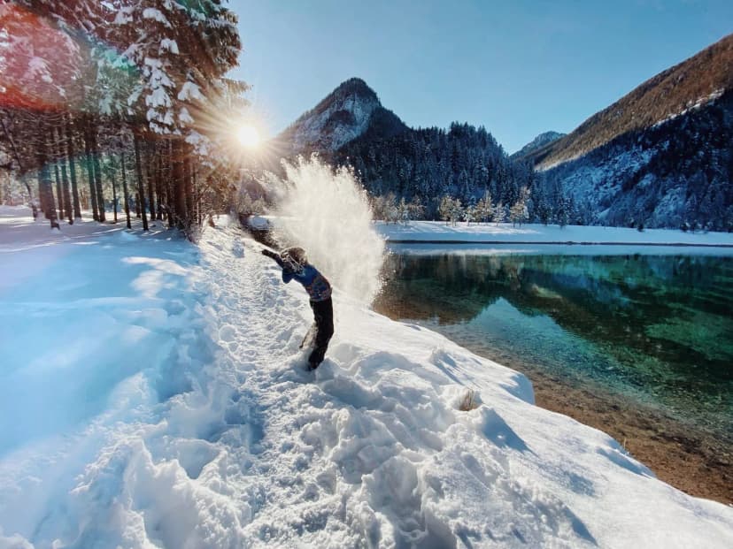 Child throwing snow by clear alpine lake with sunlit mountains in winter Slovenia