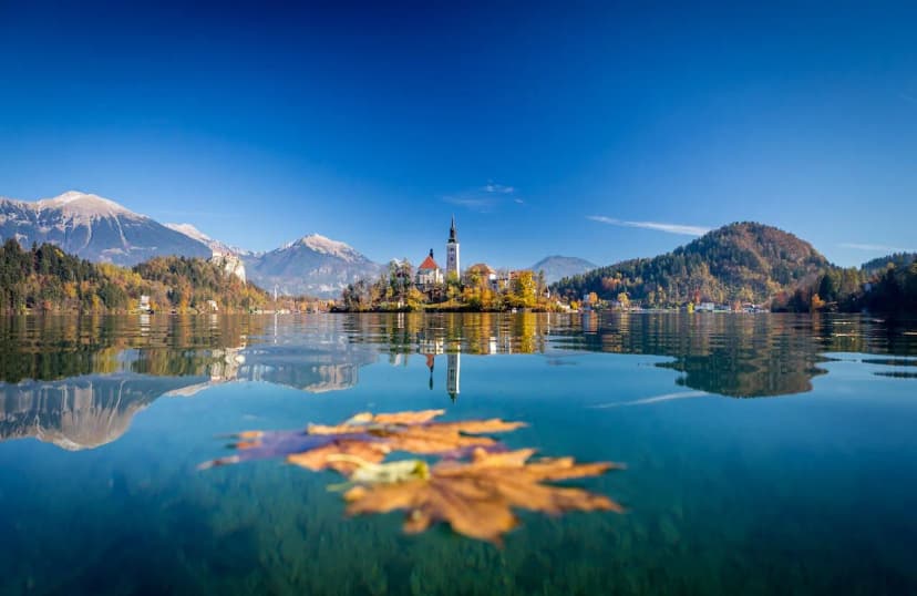 Lake Bled island church with autumn foliage, mountains, and floating maple leaf.