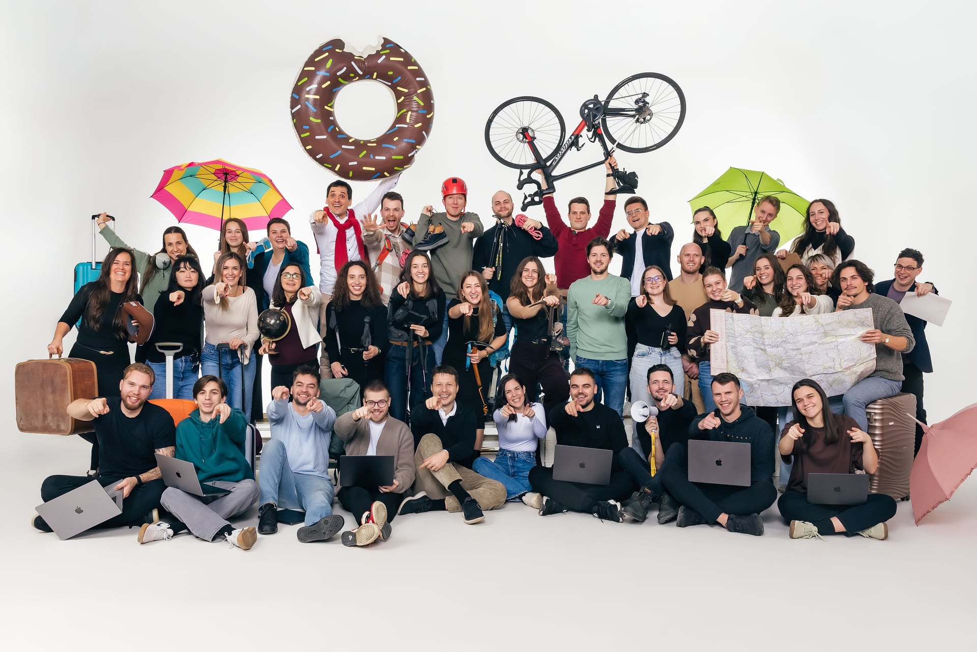 Gran grupo de personas sosteniendo artículos de viaje como una bicicleta, flotador de dona, paraguas y un mapa contra un fondo blanco.