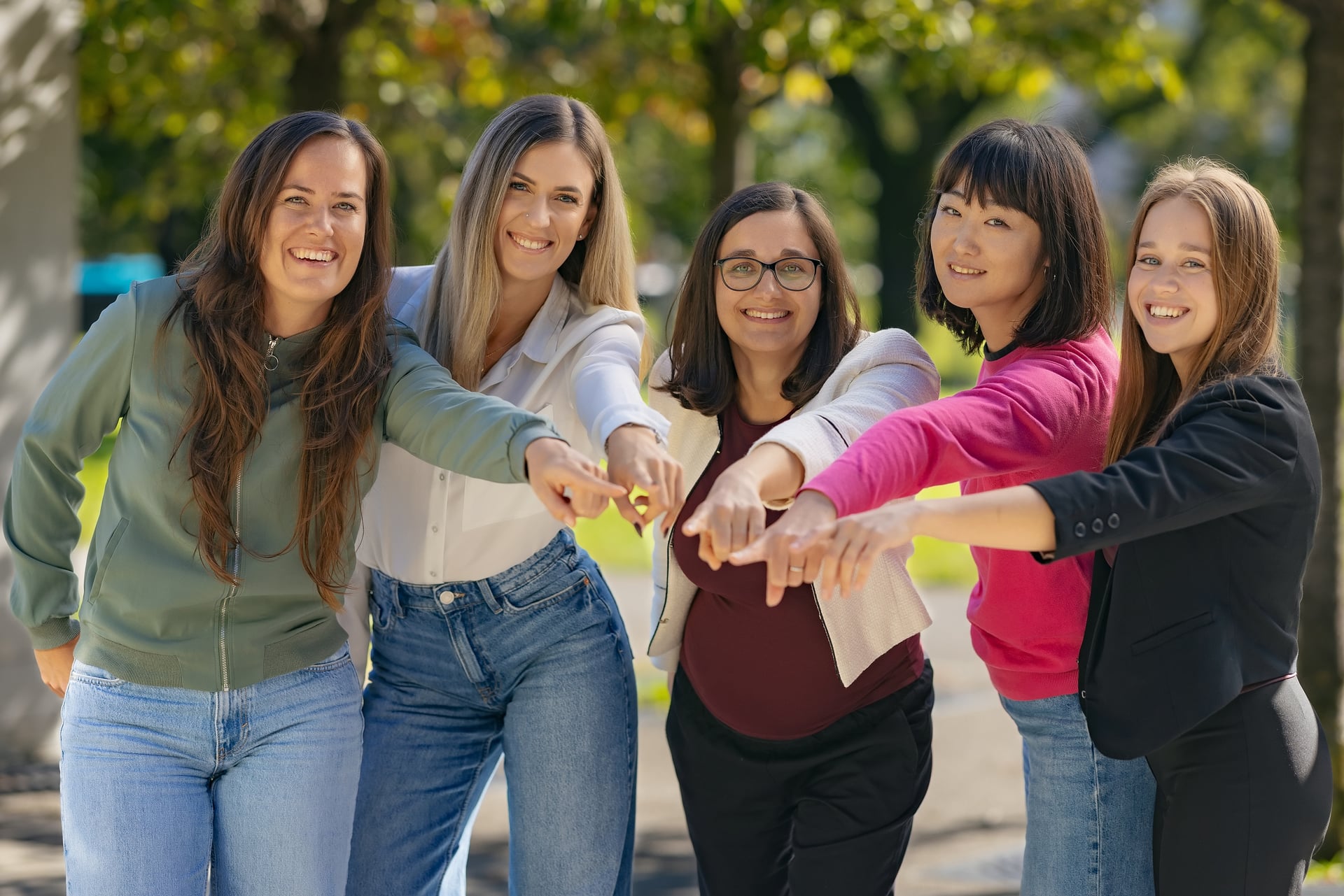 Cinco mujeres sonriendo y señalando con los dedos hacia la cámara en un parque al aire libre.