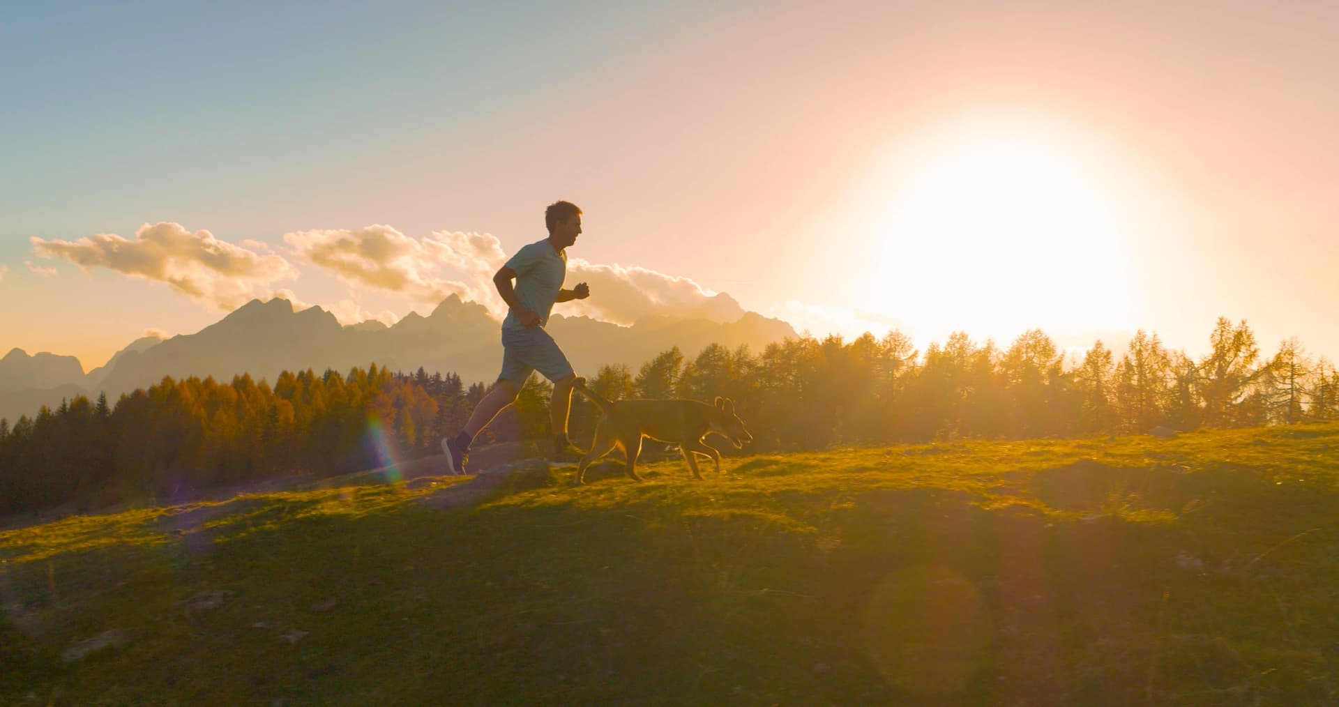 CLOSE UP, LENS FLARE: Male runner running with his dog along a scenic hilltop