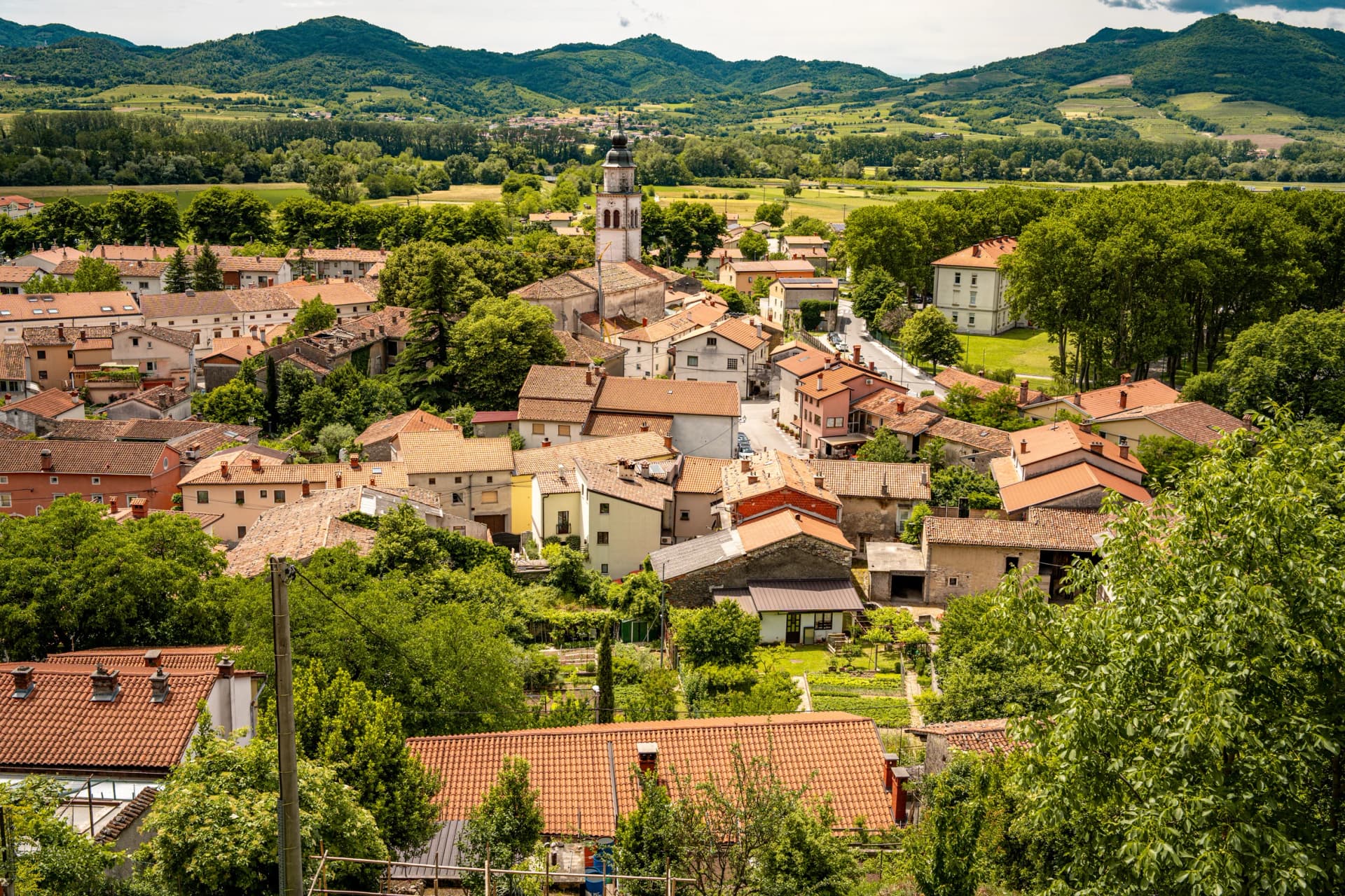 Picturesque View Of Vipava From Castle Ruins: Red-Tiled Rooftops And Church Tower Nestled In Green Valley With Blooming Flowers In Foreground And Rolling Fields In Background, Slovenia