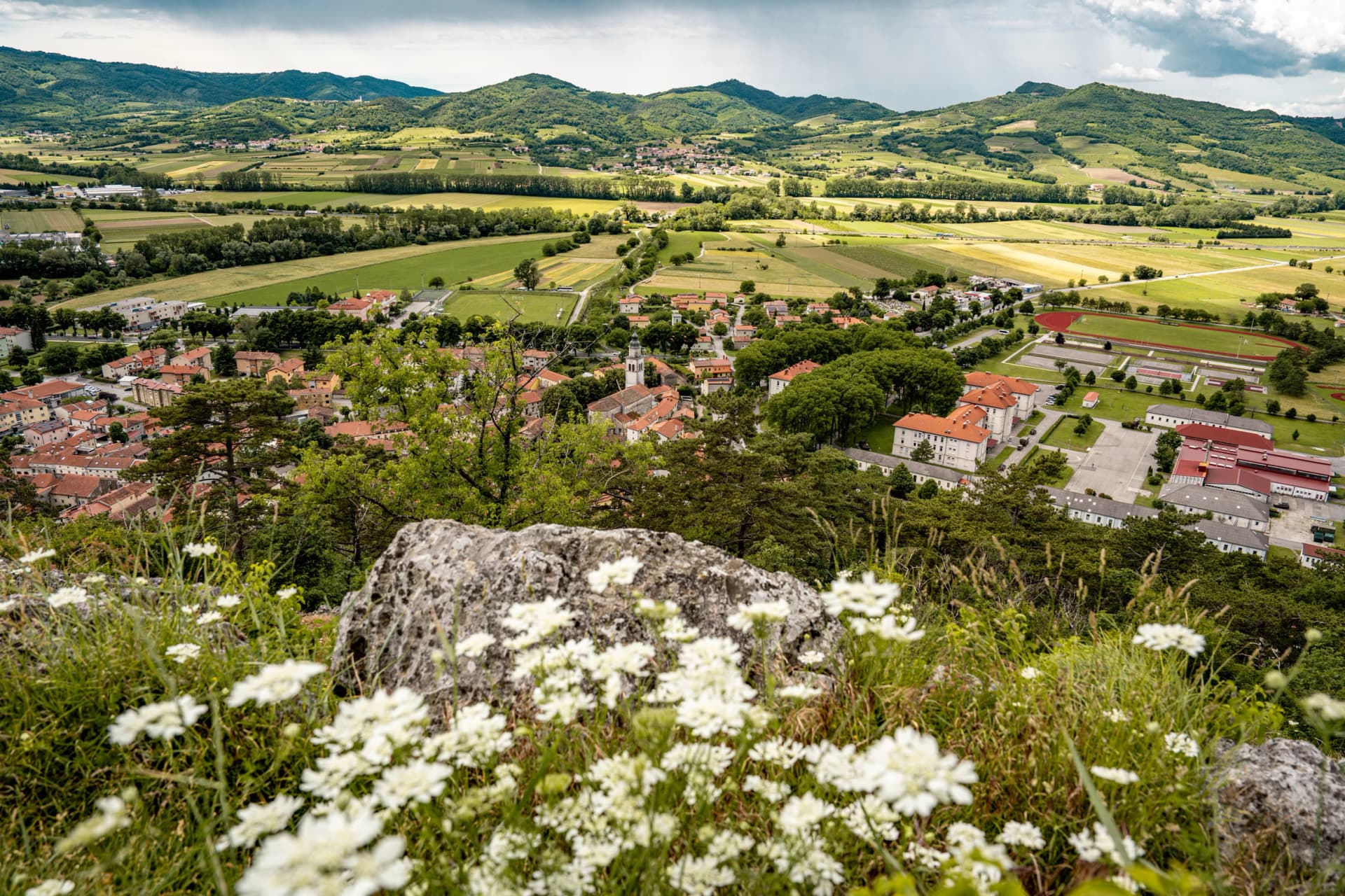 Picturesque View Of Vipava From Castle Ruins: Red-Tiled Rooftops And Church Tower Nestled In Green Valley With Blooming Flowers In Foreground And Rolling Fields In Background, Slovenia